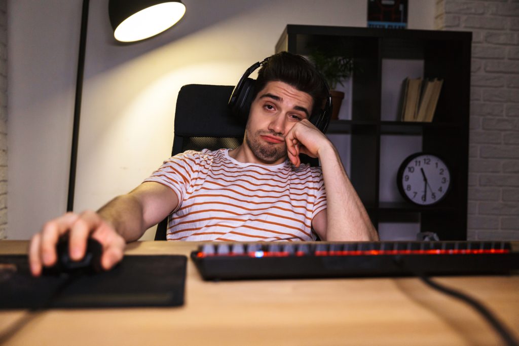 Bored gamer playing video games on computer while sitting by the table at home