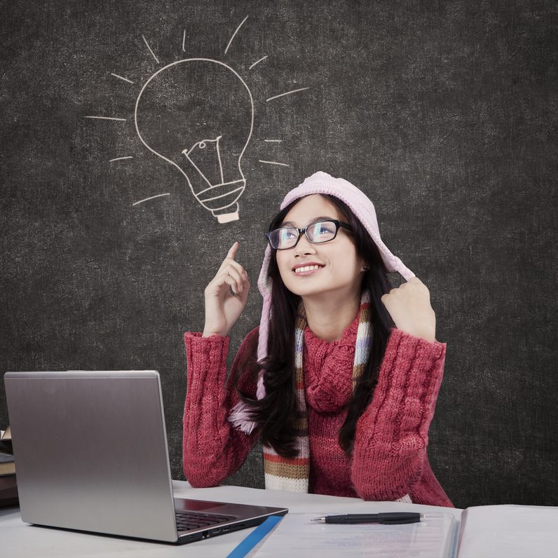 Portrait of pretty schoolgirl in knitwear studying with laptop and textbooks, pointing at lightbulb